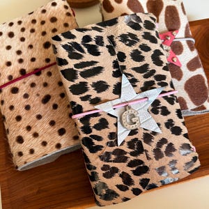 Puede incluir: Tres diarios rectangulares con cubiertas de textura animal. Uno presenta un estampado de leopardo con una estrella plateada y la letra G, otro tiene un patrón de puntos con una banda elástica rosa y el tercero tiene un estampado de vaca marrón y blanco.
