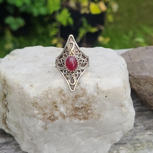 May include: A silver ring with a red gemstone set in a diamond-shaped filigree design.