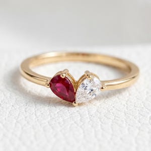 May include: A gold ring featuring two pear-shaped gemstones. One is a vibrant red, the other is clear. The ring's band is a polished gold color, and the gemstones are set close together.