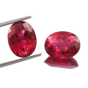May include: Two oval-cut, faceted, red gemstones. One is held by silver tweezers, the other rests on a reflective surface. The gems display a vibrant, deep red color with internal light reflections, showcasing their clarity and cut.