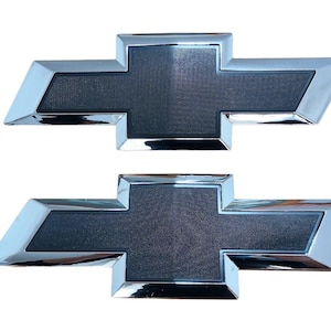 May include: Two Chevrolet bow tie emblems. Each emblem features a dark grey textured centre with a chrome-coloured border. The emblems are displayed against a white background.