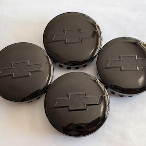 May include: Four black hubcaps with the Chevrolet logo in the center. The hubcaps are round with a glossy finish and a raised emblem. The background is a plain white surface.