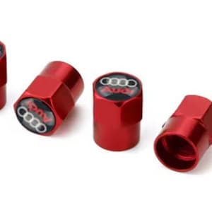 May include: Four red tire valve stem caps with the Audi logo in white and black. The caps are hexagonal and have a glossy finish. The Audi logo is centered on the top of each cap.