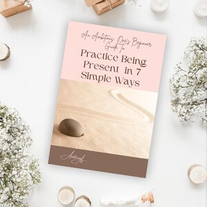 Peut inclure: Une couverture de livre avec le titre "An Ambitious One's Beginner Guide to Practice Being Present in 7 Simple Ways". La couverture présente un schéma de couleurs rose clair et beige avec un design de jardin zen et une pierre lisse et brune.
