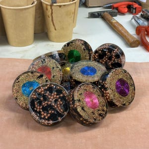 May include: A collection of handcrafted, round resin art pieces. Each piece features a central spiral design in various colors, including blue, green, red, and pink, surrounded by gold-colored wire and embedded with dark, textured elements. The pieces are displayed on a light brown surface.