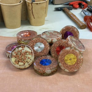 Peut inclure: Plusieurs pièces d'art en résine transparentes et rondes, avec du fil de cuivre et de petites pierres. Certaines pièces présentent un motif central en spirale argenté, rouge, bleu ou jaune. Les pièces sont exposées sur une surface brun clair.