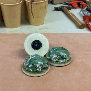 May include: Three decorative resin objects. Two are dome-shaped with green and silver inclusions, and one is a cylindrical shape with a black center. The objects are on a light brown surface.