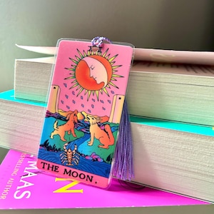 May include: A pink tarot card bookmark featuring "The Moon" card illustration. The design includes a sun with a crescent moon, two dogs, a river, and a scorpion. A purple tassel and cord are attached to the top. The bookmark is resting on a stack of books.