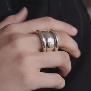 Op de afbeelding: Een zilveren ring met een afgerond middenstuk en twee gebogen banden aan weerszijden. De ring wordt aan een vinger gedragen, waarbij het gladde, reflecterende oppervlak wordt benadrukt. Het sieraad heeft een modern, minimalistisch ontwerp.