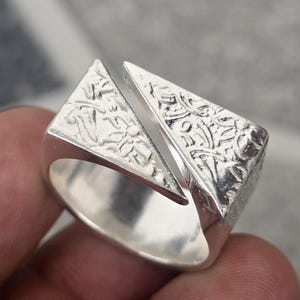 May include: A silver ring with a geometric design. The ring features two textured, triangular shapes that are separated by a small gap. The ring has a smooth, polished band.