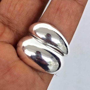 May include: A shiny, silver ring with a unique design. The ring features two smooth, rounded shapes that appear to be connected. The ring is worn on a finger, and the polished surface reflects light.
