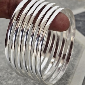 May include: A collection of shiny, silver bangles. The bracelets are smooth and reflective, with a rounded, flat profile. Light reflects off the polished metal, creating highlights and shadows. The bangles are stacked together, creating a layered effect.