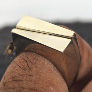 以下が含まれることがあります： 長方形の面を持つ、金色の幾何学的なリング。 リングはモダンで角張ったデザインで、研磨仕上げです。 リングは人の指に表示されています。