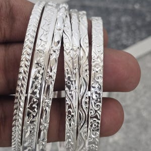 May include: A selection of silver bangles, each adorned with detailed floral and leaf patterns. The bangles are presented, highlighting their intricate designs and polished finish. A classic jewellery accessory.