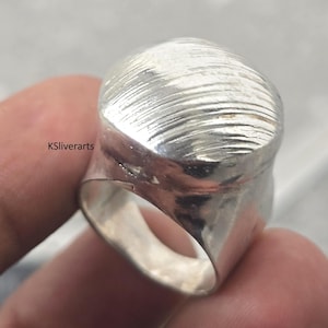 Puede incluir: Primer plano de un anillo de plata con una parte superior texturizada y abovedada. El anillo tiene una banda ancha y un acabado liso y pulido en los laterales. La parte superior del anillo tiene una serie de líneas horizontales. El texto "KSliverarts" es visible.