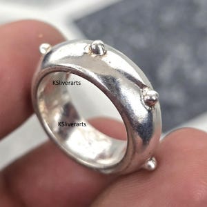 May include: A silver ring with a rounded band and small, raised spherical details. The ring has a polished finish and appears to be made of metal. The text "KSliverarts" is visible on the ring.