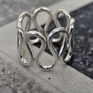 May include: A silver ring with a distinctive design of interconnected figure-eight shapes. The ring is made of a shiny, reflective metal and is set against a neutral background. The text "KSliverarts" is visible on the ring.