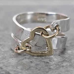 Peut inclure: Bague en argent avec un charm en forme de cœur doré. La bague présente un design à deux bandes, avec un charm en forme de cœur doré reliant les bandes. La bague est sur une surface grise.