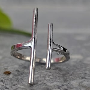 May include: A silver-colored ring with a modern design. The ring features two vertical bars that extend upwards from the band. The ring is open, allowing for some adjustment in size. The brand name "KSliverarts" is visible on the ring.