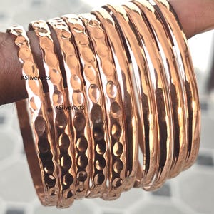 Peut inclure: Plusieurs bracelets en cuivre, certains avec une texture martelée et d'autres avec une finition lisse. Les bracelets sont présentés sur une main, mettant en valeur leur couleur métallique chaude. Le texte "KSliverarts" est visible.