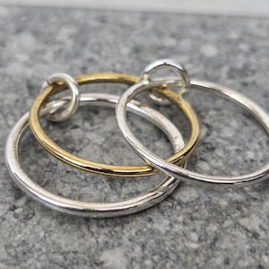 May include: Three interlocking rings, two silver and one gold, on a textured grey surface. The rings are smooth and circular, with a simple, elegant design. The gold ring adds warmth to the composition.