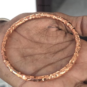 Op de afbeelding: Een gehamerde koperen armband. De armband heeft een getextureerd oppervlak met een gehamerd ontwerp. Het koper heeft een warme, roodbruine kleur. De armband is cirkelvormig en lijkt handgemaakt.