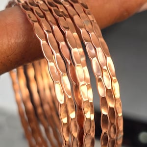 Puede incluir: Primer plano de varias pulseras de color cobre con un patrón geométrico texturizado. Las pulseras están apiladas, reflejando la luz y creando un brillo metálico cálido. La joyería es un accesorio simple pero elegante.