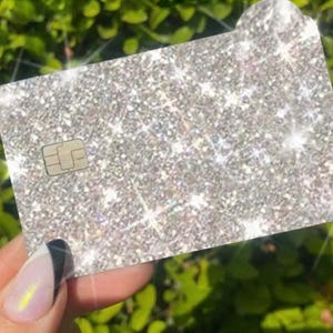 Puede incluir: Una tarjeta de cr&eacute;dito plateada con purpurina, con la parte superior redondeada y un microchip. La tarjeta se sostiene delante de un fondo de hojas verdes. La tarjeta tiene un aspecto brillante y brillante.