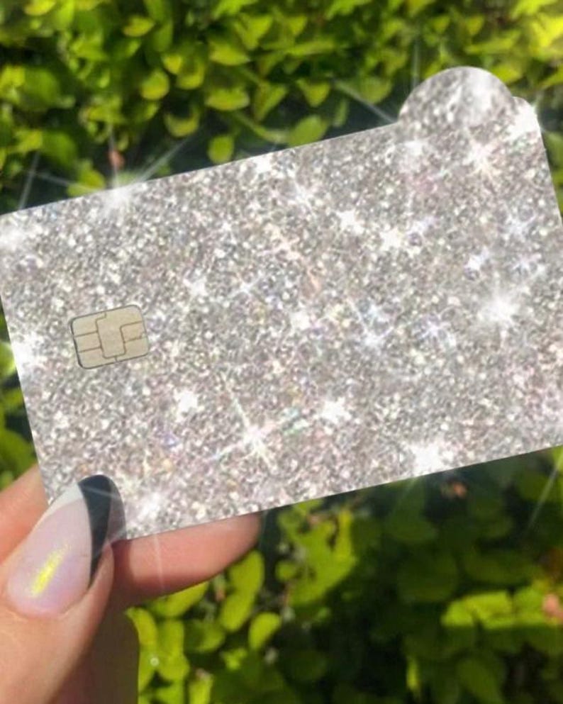 Puede incluir: Una tarjeta de cr&eacute;dito plateada con purpurina, con la parte superior redondeada y un microchip. La tarjeta se sostiene delante de un fondo de hojas verdes. La tarjeta tiene un aspecto brillante y brillante.