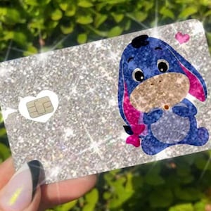 May include: A glittery silver card featuring a cartoon image of Eeyore from Winnie the Pooh. The blue Eeyore has pink ears and a pink bow. A small heart and a white heart with a chip are also on the card.