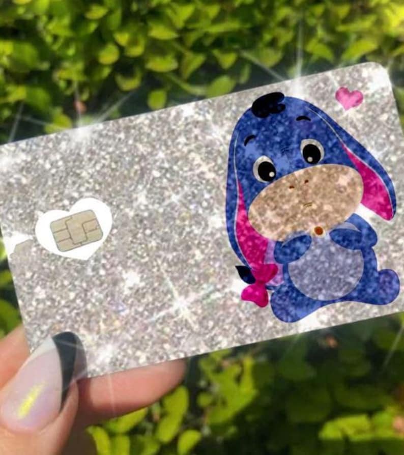 May include: A glittery silver card featuring a cartoon image of Eeyore from Winnie the Pooh. The blue Eeyore has pink ears and a pink bow. A small heart and a white heart with a chip are also on the card.