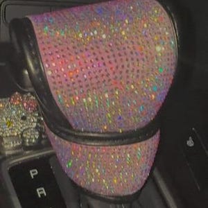 May include: A car gear shift knob cover featuring a black leather-like trim and a surface densely covered in iridescent, multi-colored rhinestones that reflect light. A small, rhinestone-encrusted character emblem is visible to the left side.