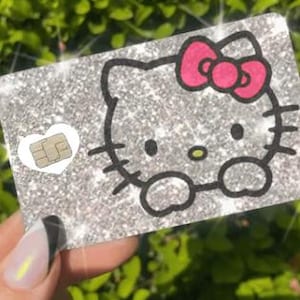 Op de afbeelding: Een glinsterende zilveren kaart met een Hello Kitty-ontwerp met een roze strik en zwarte omtrek. Een klein wit hart met een chip bevindt zich aan de linkerkant. De kaart wordt tegen een vage groene achtergrond gehouden.