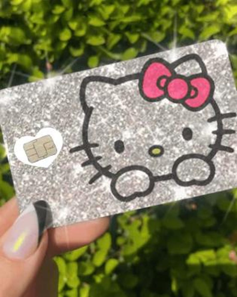 Op de afbeelding: Een glinsterende zilveren kaart met een Hello Kitty-ontwerp met een roze strik en zwarte omtrek. Een klein wit hart met een chip bevindt zich aan de linkerkant. De kaart wordt tegen een vage groene achtergrond gehouden.