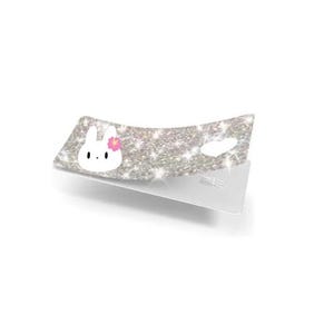 May include: A sparkly silver accessory with a white bunny face and pink flower. The accessory is on a white stand, and the surface is covered in glitter.