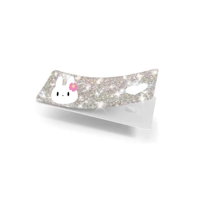 May include: A sparkly silver accessory with a white bunny face and pink flower. The accessory is on a white stand, and the surface is covered in glitter.
