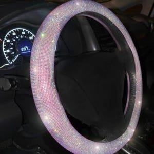 May include: A sparkling steering wheel cover, encrusted with iridescent rhinestones. The cover is a light pink color and is designed to fit over a car's steering wheel. The dashboard and other car components are visible in the background.