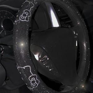 May include: Black steering wheel cover embellished with sparkling rhinestones. The cover features a repeating pattern of a cartoon character. The steering wheel is black and appears to be in a car.