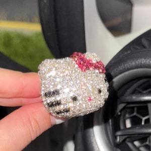 May include: A decorative accessory shaped like a cat head, fully encrusted with clear, sparkling rhinestones. It features a bright pink rhinestone bow on the top right, black rhinestone whiskers on the left side, and small black rhinestone eyes.