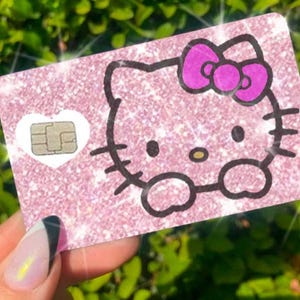 May include: A pink glittery card featuring a Hello Kitty design with a purple bow. The card has a white heart-shaped chip and is held against a backdrop of green foliage.