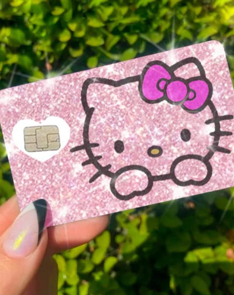 May include: A pink glittery card featuring a Hello Kitty design with a purple bow. The card has a white heart-shaped chip and is held against a backdrop of green foliage.