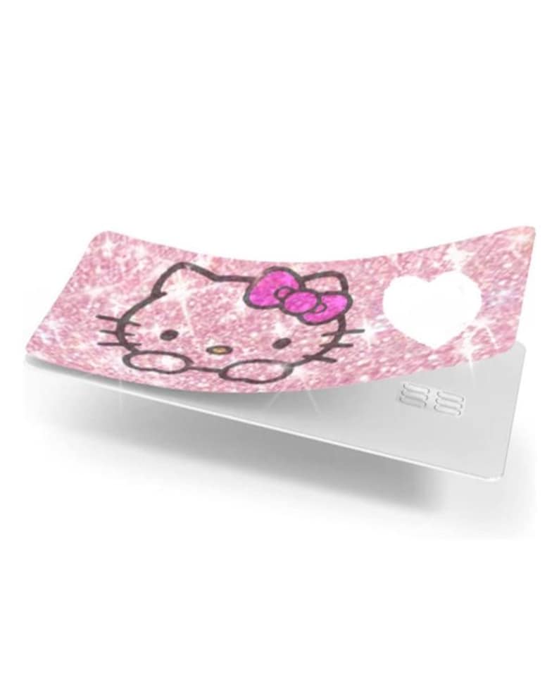 May include: A pink glittery card with a Hello Kitty design and a white heart cutout. The Hello Kitty graphic has a pink bow and is outlined in black. The card is on a white surface.