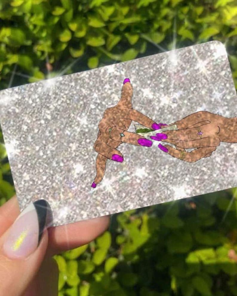 Może przedstawiać: Błyszczący srebrny prostokąt z ilustracją dłoni zwijających jointa. Dłonie mają fioletowe paznokcie i złote pierścionki. Tło jest rozmyte zielone.