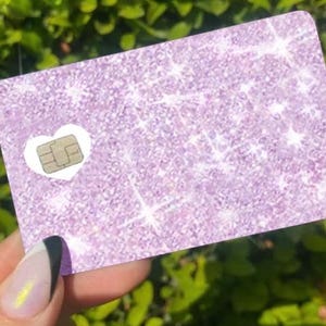 May include: A rectangular card with a glittery lavender surface. A white heart shape surrounds a small, gold-colored chip. The card is held in front of a blurred green background.