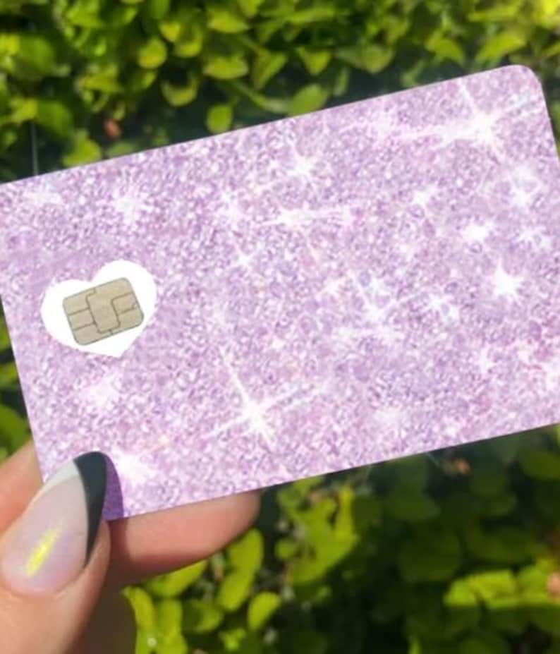 May include: A rectangular card with a glittery lavender surface. A white heart shape surrounds a small, gold-colored chip. The card is held in front of a blurred green background.