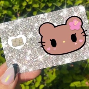 以下が含まれることがあります： キラキラ光る銀色のカード。漫画のクマの顔、ピンクの花、ハート型のほお紅が描かれています。カードには金のチップが付いた白いハートがあります。カードは、マニキュアされた爪のある手で持たれています。