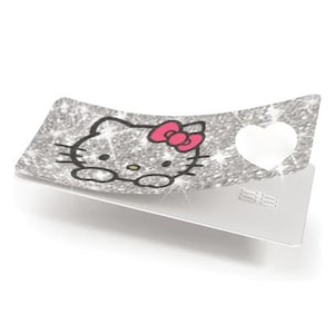 Op de afbeelding: Een zilveren glitterkaart met een Hello Kitty-ontwerp. Het ontwerp toont het gezicht van Hello Kitty met een roze strik. Een witte hartvorm is uitgesneden aan de rechterkant van de kaart. De onderkant van de kaart is zilver.