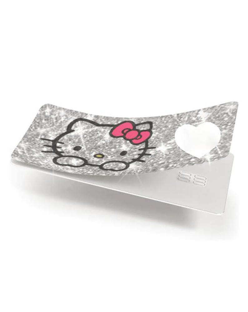 Op de afbeelding: Een zilveren glitterkaart met een Hello Kitty-ontwerp. Het ontwerp toont het gezicht van Hello Kitty met een roze strik. Een witte hartvorm is uitgesneden aan de rechterkant van de kaart. De onderkant van de kaart is zilver.