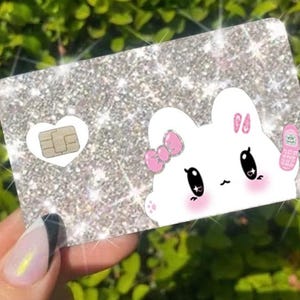 K&ouml;nnte beinhalten: Glitzernde silberne Karte mit einem Cartoon-Hasen-Design. Der Hase hat eine rosa Schleife, Rouge und ein kleines rosa Telefon. Eine wei&szlig;e Herzform enth&auml;lt eine SIM-Karte. Die Karte wird vor einem gr&uuml;nen, bl&auml;ttrigen Hintergrund gehalten.