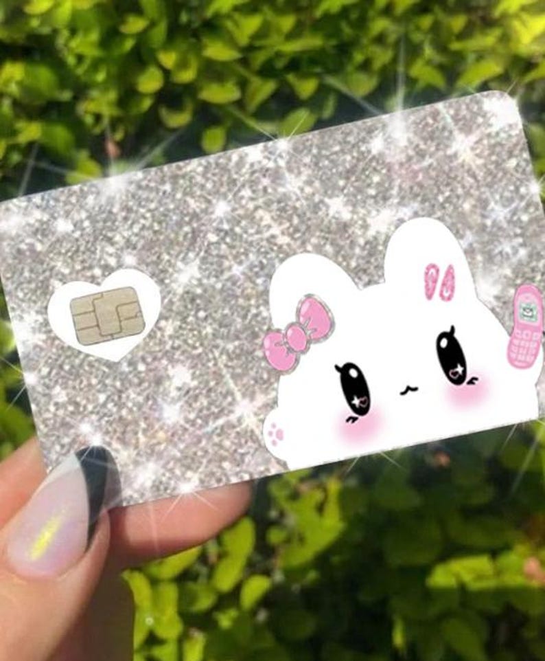 K&ouml;nnte beinhalten: Glitzernde silberne Karte mit einem Cartoon-Hasen-Design. Der Hase hat eine rosa Schleife, Rouge und ein kleines rosa Telefon. Eine wei&szlig;e Herzform enth&auml;lt eine SIM-Karte. Die Karte wird vor einem gr&uuml;nen, bl&auml;ttrigen Hintergrund gehalten.